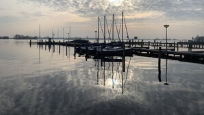 Ein ruhiger Yachthafen mit vertäuten Booten, einer anlegenden Pier und einem wolkenlosen Himmel, in dem die Sonne zu sehen ist.