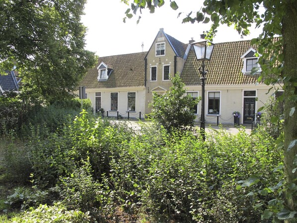 A traditional building with a tiled roof, a central entrance, and a small garden area with dense foliage in the foreground. - De Thuiskamer Just like at home! (Grou)