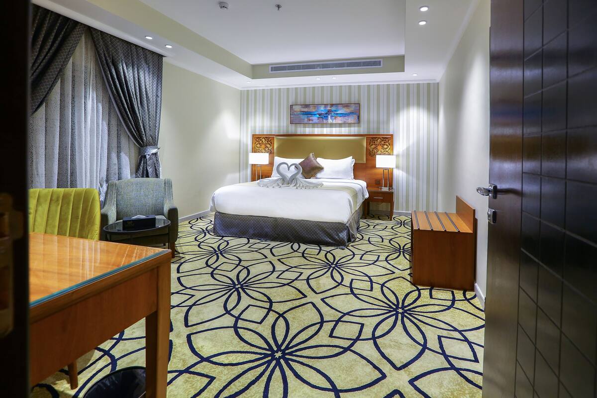 A hotel room with a large bed, two bedside tables with lamps, a small table with a chair, a decorative pillow, and a patterned carpet.