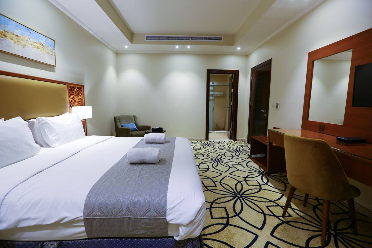 A hotel room with a large bed, a desk, a chair, a mirror, and a bathroom visible through an open door.