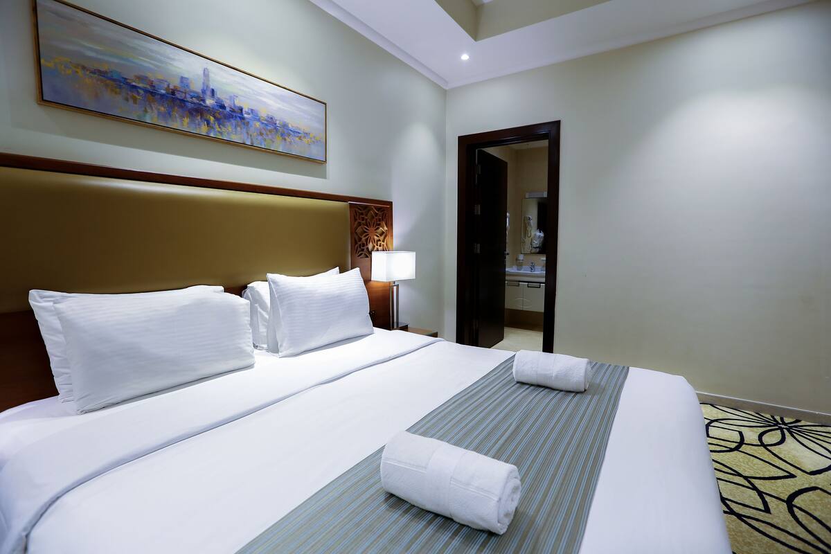 A hotel room with a large bed, a headboard, a painting, a nightstand, and a lamp.