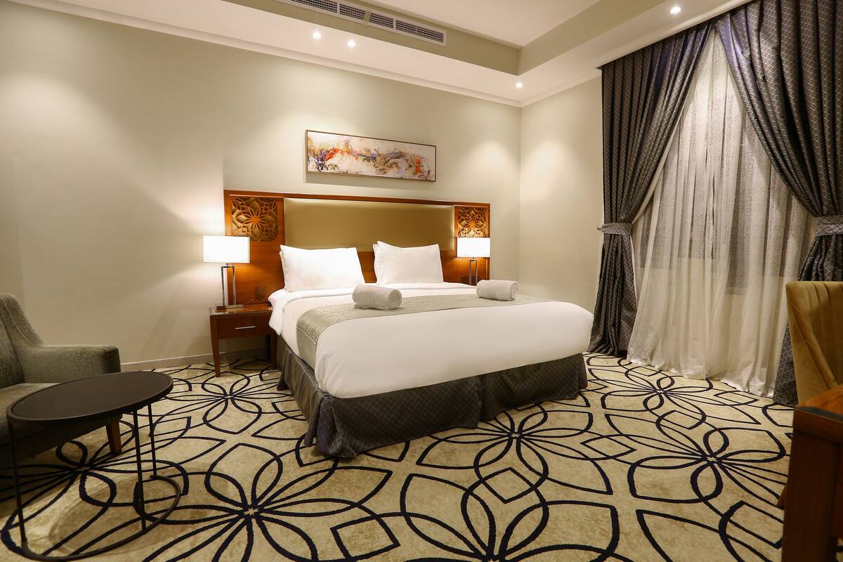 A hotel room with a large bed, two bedside tables with lamps, a chair, and a decorative headboard.