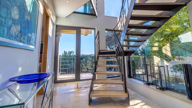 Un interior moderno con una escalera, barandas de vidrio y vistas al exterior.