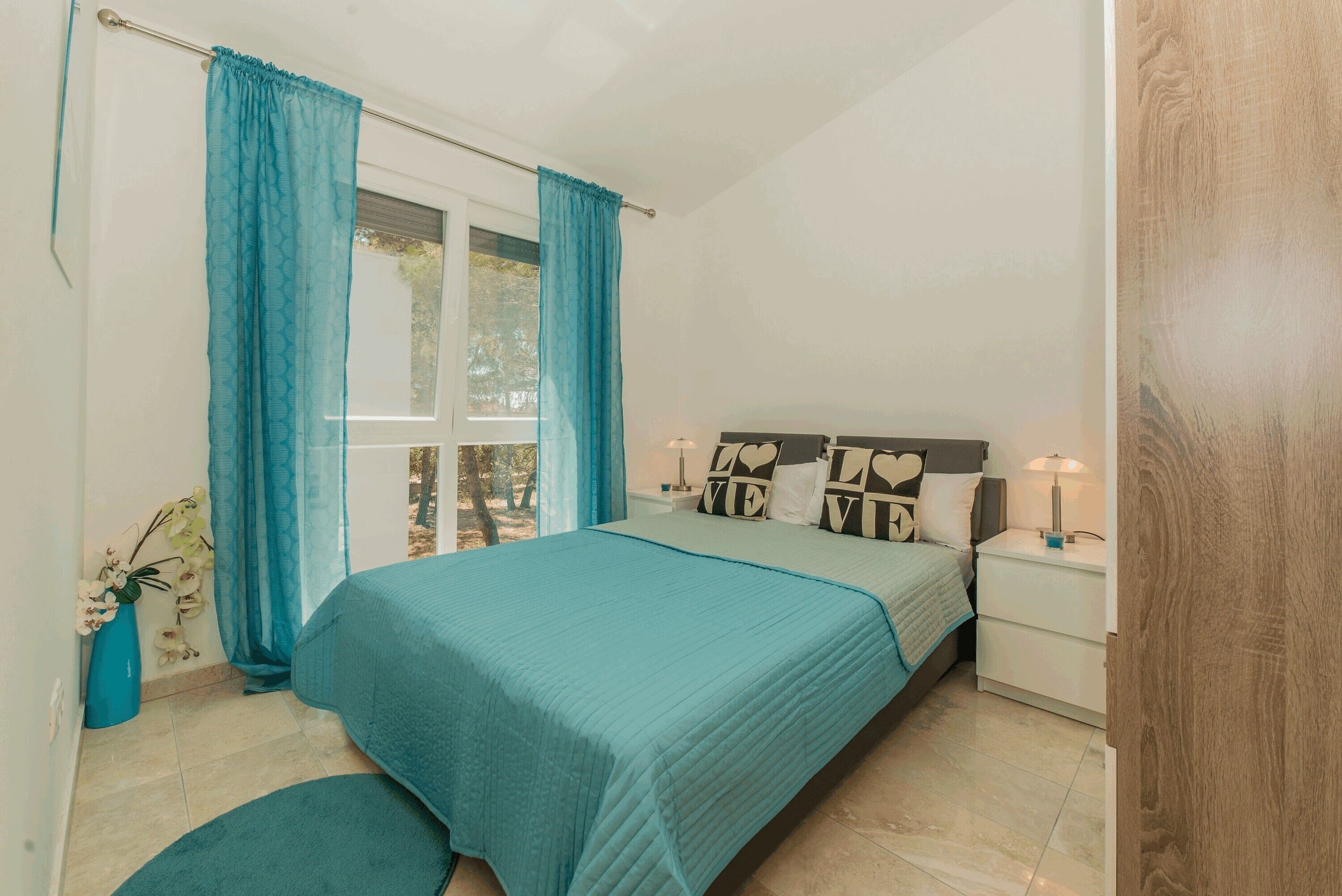 A bedroom with a bed, a nightstand, a window with curtains, and a plant.