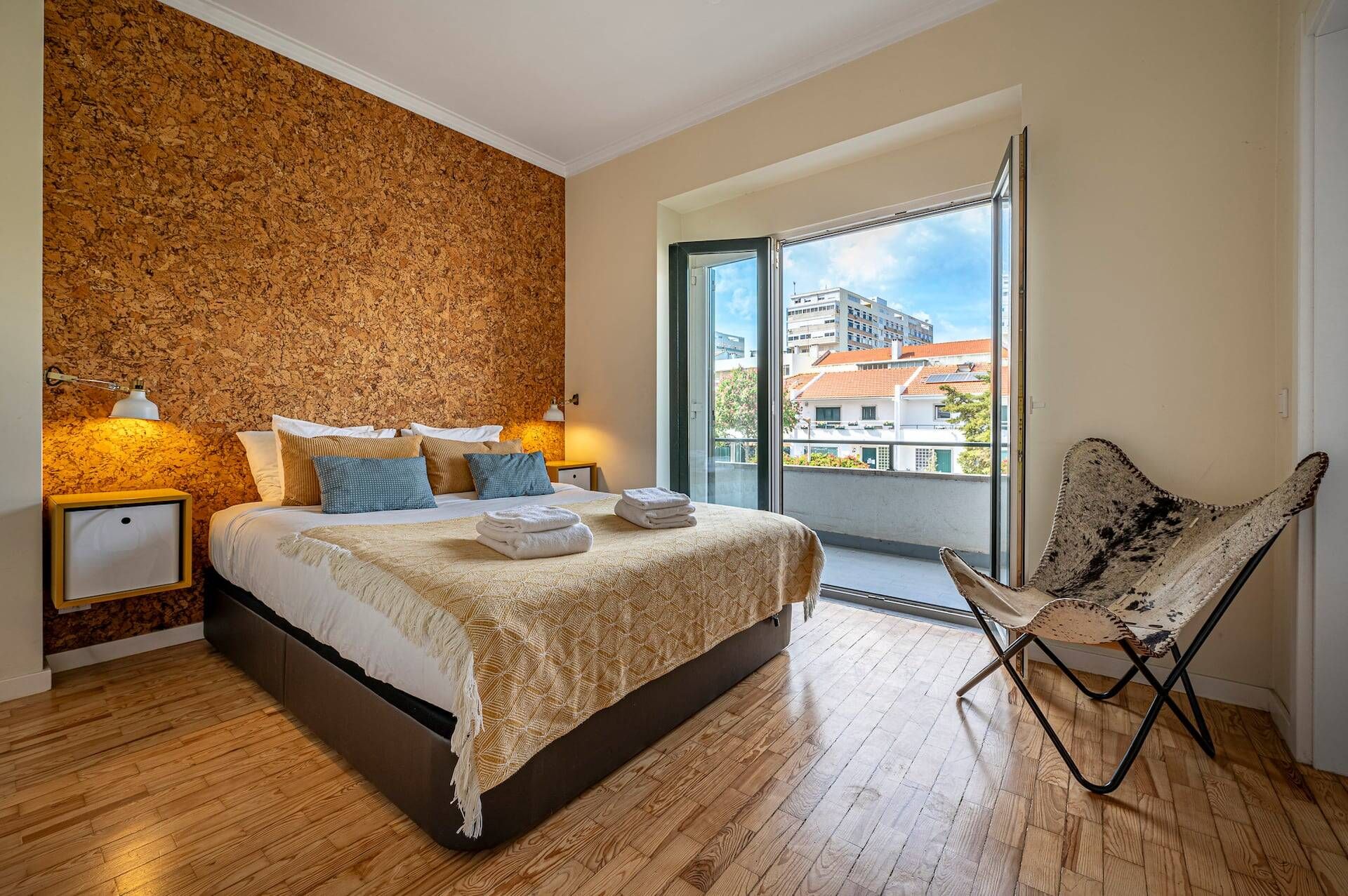 A modern bedroom with a large bed, a wooden floor, a balcony with a view of buildings, and a chair.