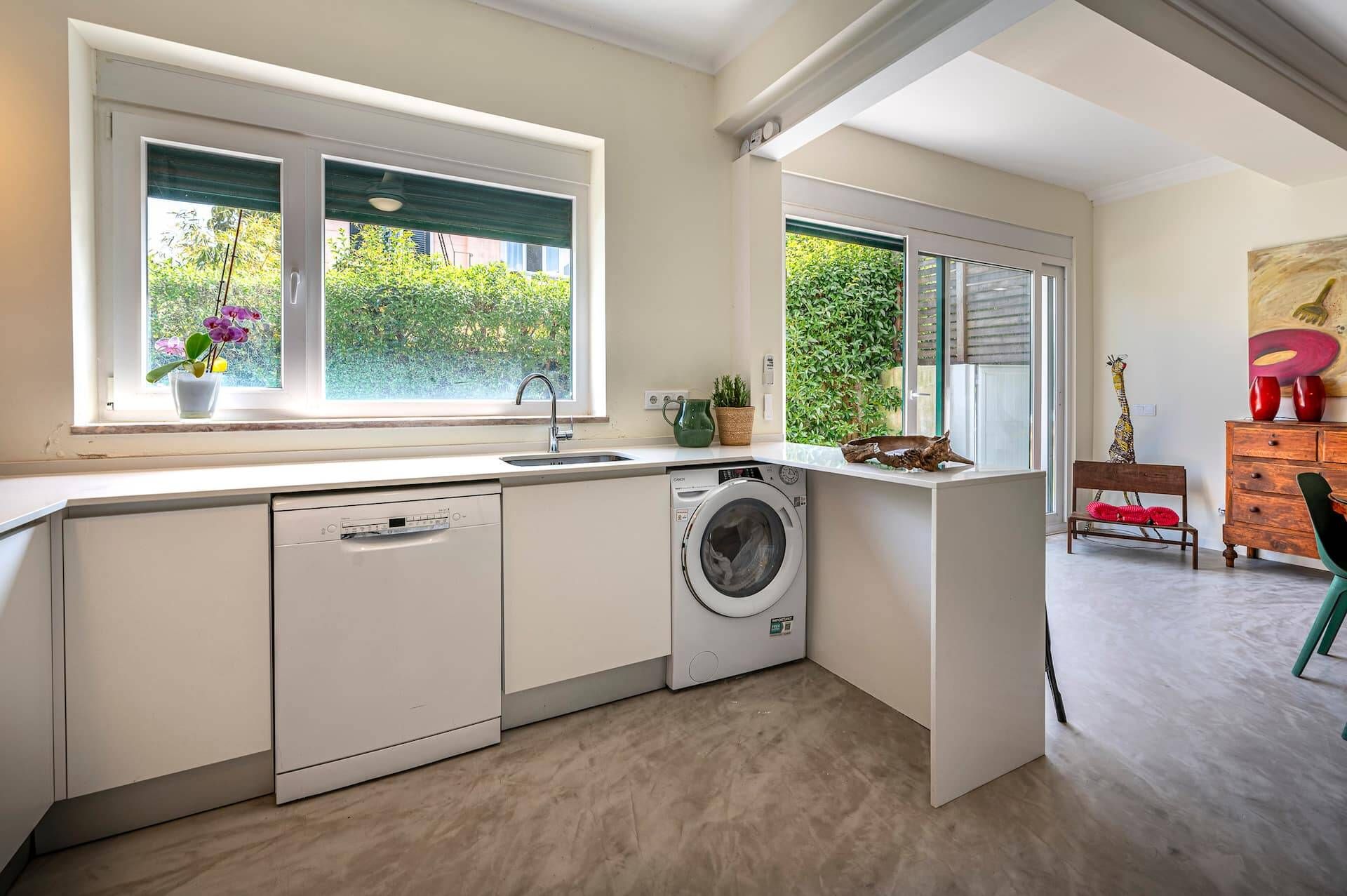A compact kitchen with a dishwasher, washing machine, and a window with a view of greenery.