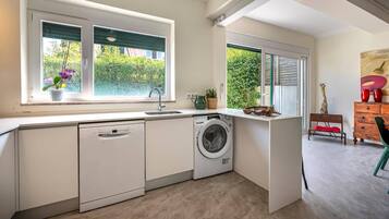 A compact kitchen with a dishwasher, washing machine, and a window with a view of greenery.