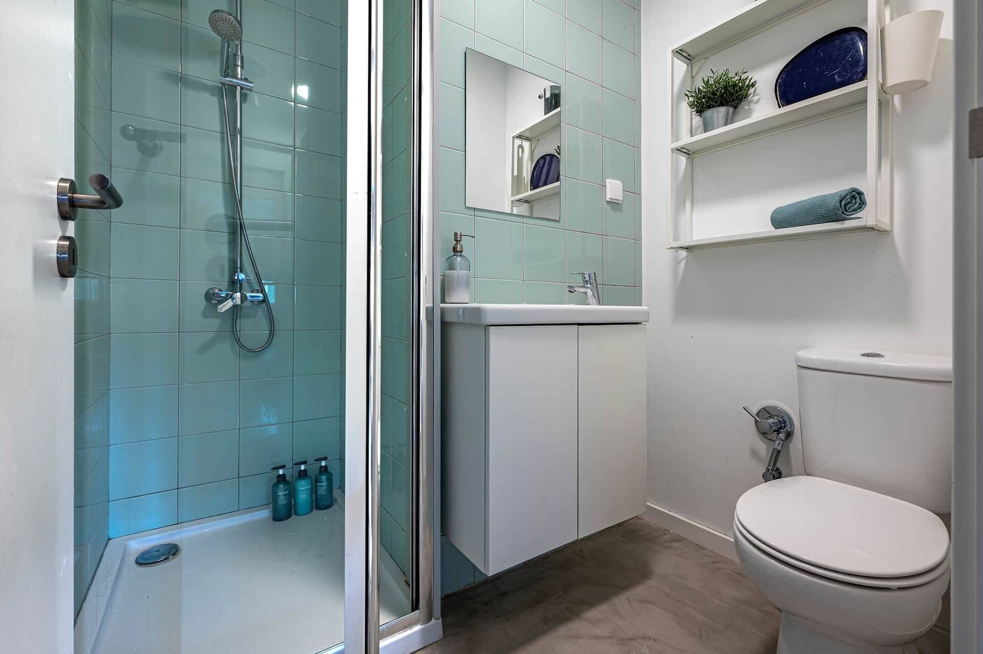 A bathroom with a glass-enclosed shower, a white toilet, a sink with a mirror above it, and a shelf with toiletries.