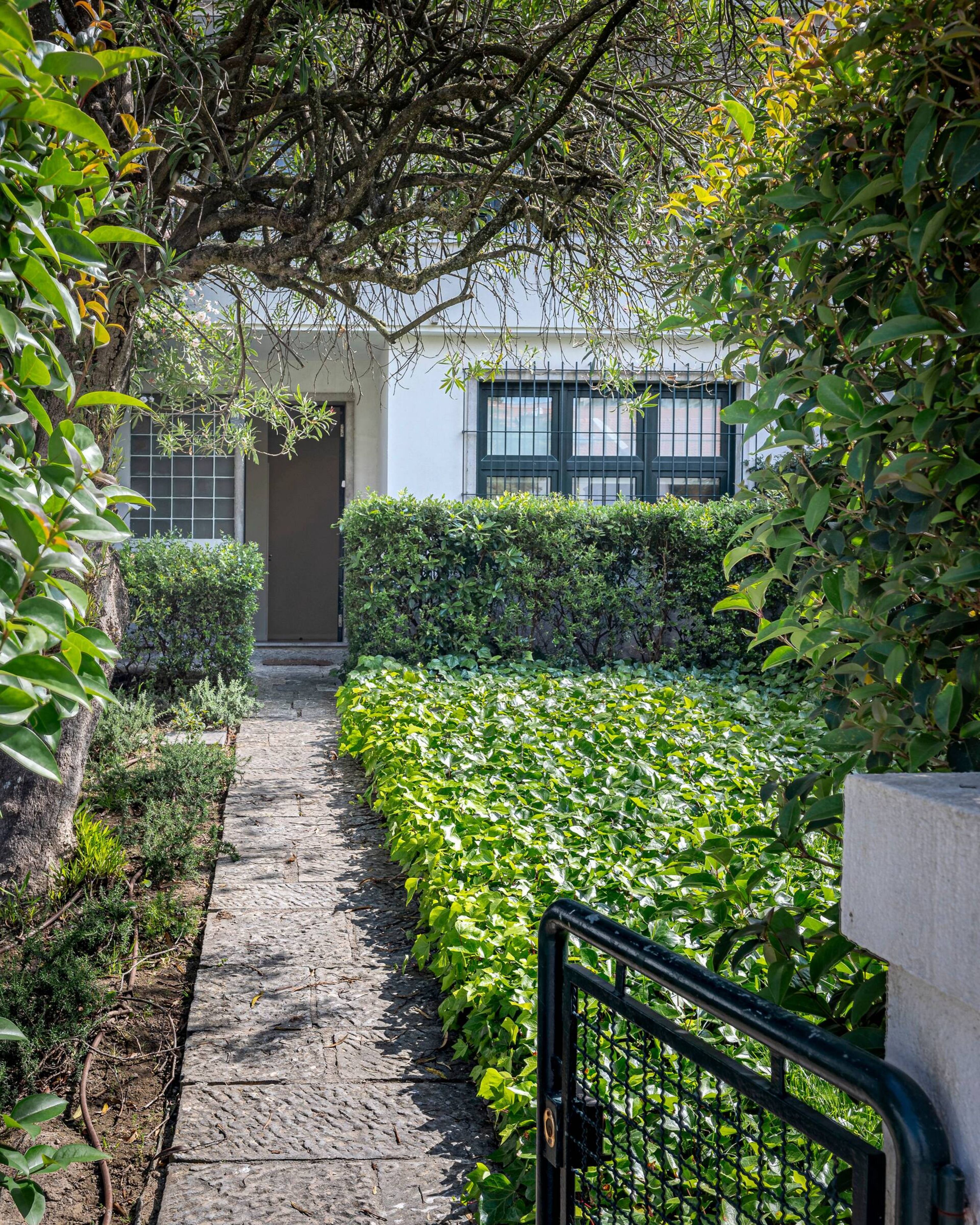 A stone pathway leading to a building entrance, surrounded by lush greenery and a metal gate.