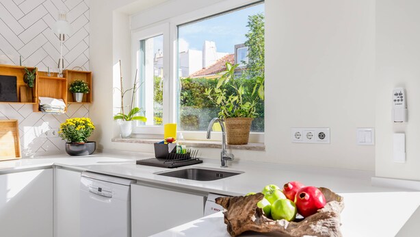 Eine moderne Küche mit großem Fenster, weißen Schränken und einer Obstschale auf der Arbeitsfläche.