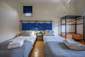 A bedroom with two beds, a nightstand, a lamp, a window with a blind, and a wooden shelf with hangers and crates.