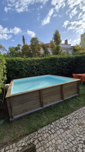 A rectangular wooden swimming pool with a stone border, surrounded by a hedge and a stone pathway.