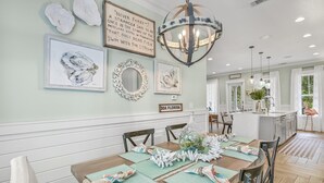 A dining area with a wooden table set for four, surrounded by chairs. There are wall-mounted artworks, a chandelier, and a sign indicating the room number. - Renovated Seagrove Home with LSV & Pool Access (Santa Rosa Beach)