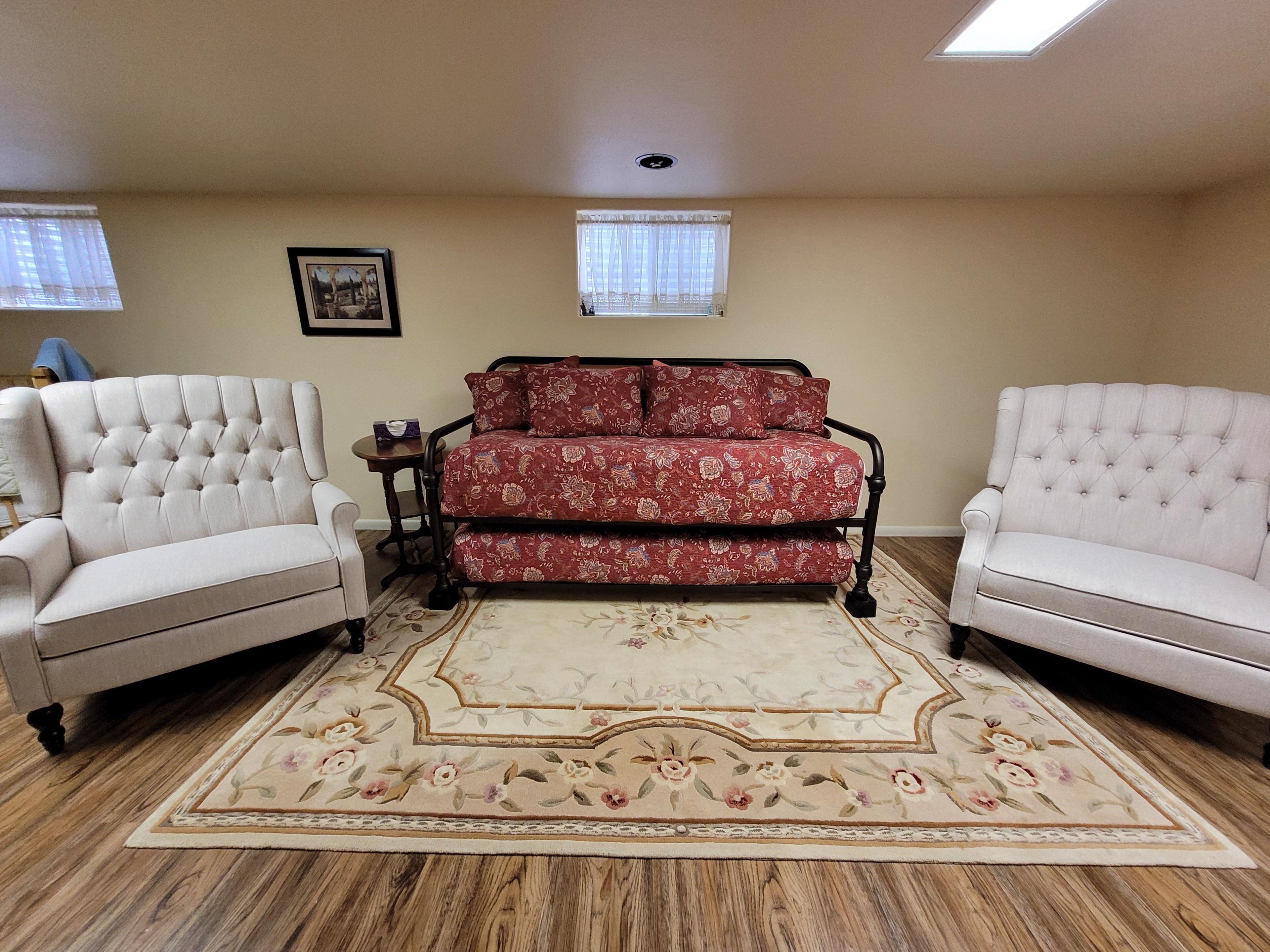 Une pièce comprenant deux fauteuils blancs, un canapé rouge à motifs, un tapis à fleurs et un tableau encadré accroché au mur.