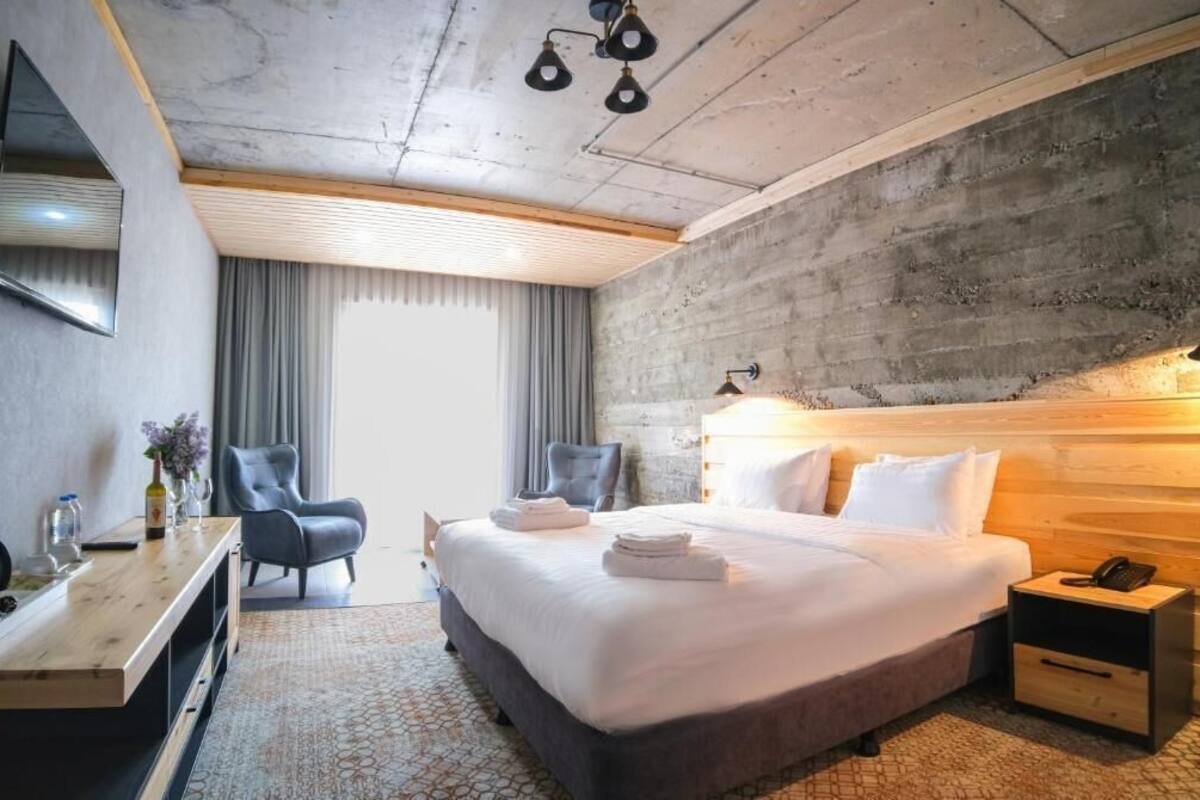 A modern hotel room with a large bed, a desk, a chair, and a view of the outdoors.