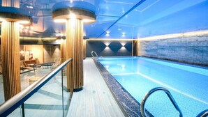 Una piscina interior con plataforma de madera y barandillas de vidrio, rodeada de columnas de madera y un diseño de techo moderno.