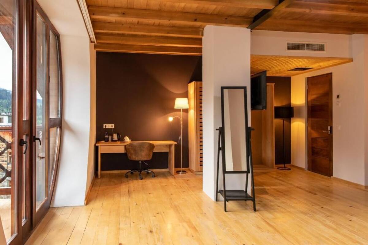 A room with wooden flooring, a desk with a chair, a standing lamp, and a TV mounted on the wall.