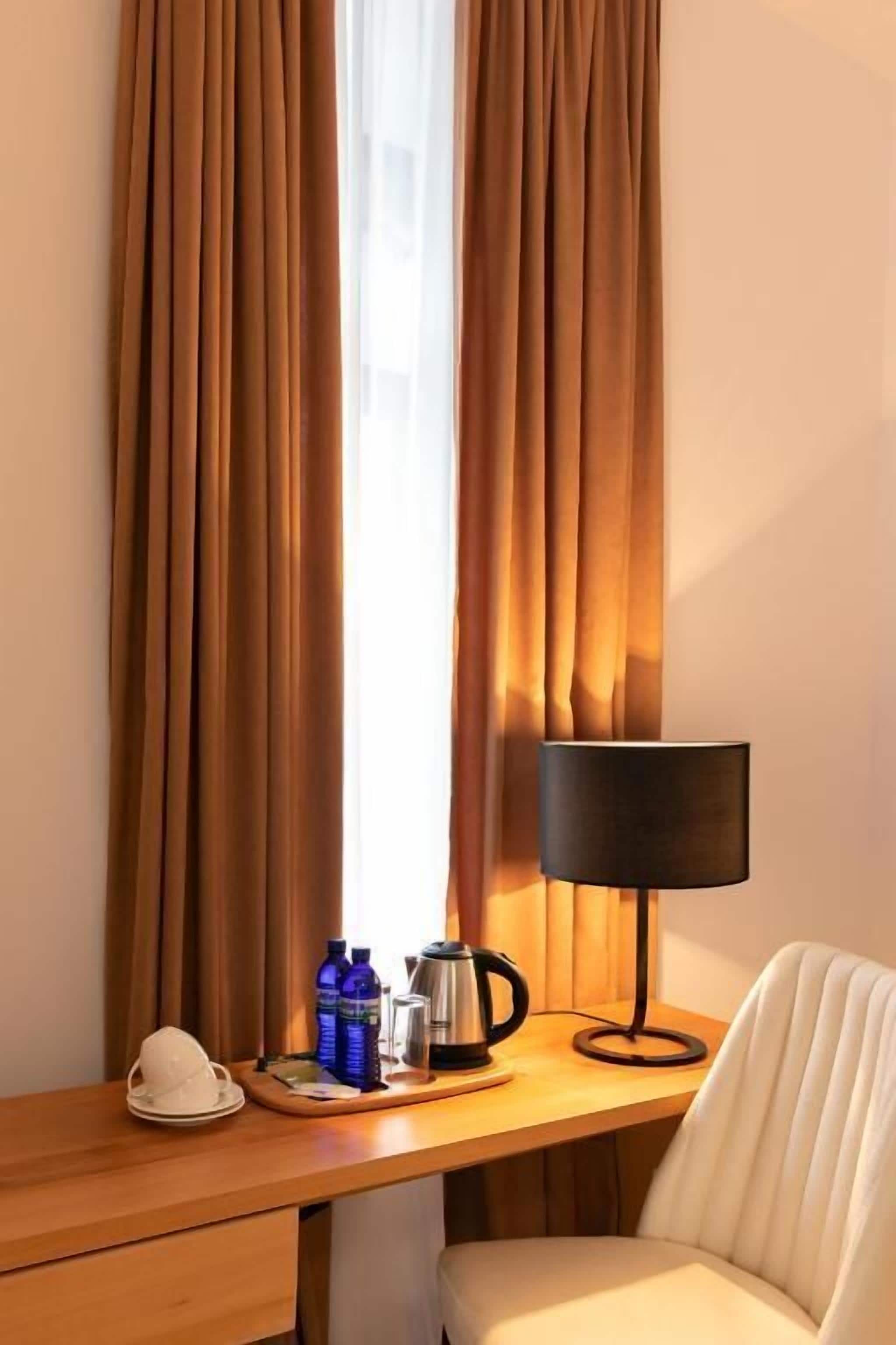 A hotel room with a wooden desk, a white chair, a black lamp, and a window with brown curtains.