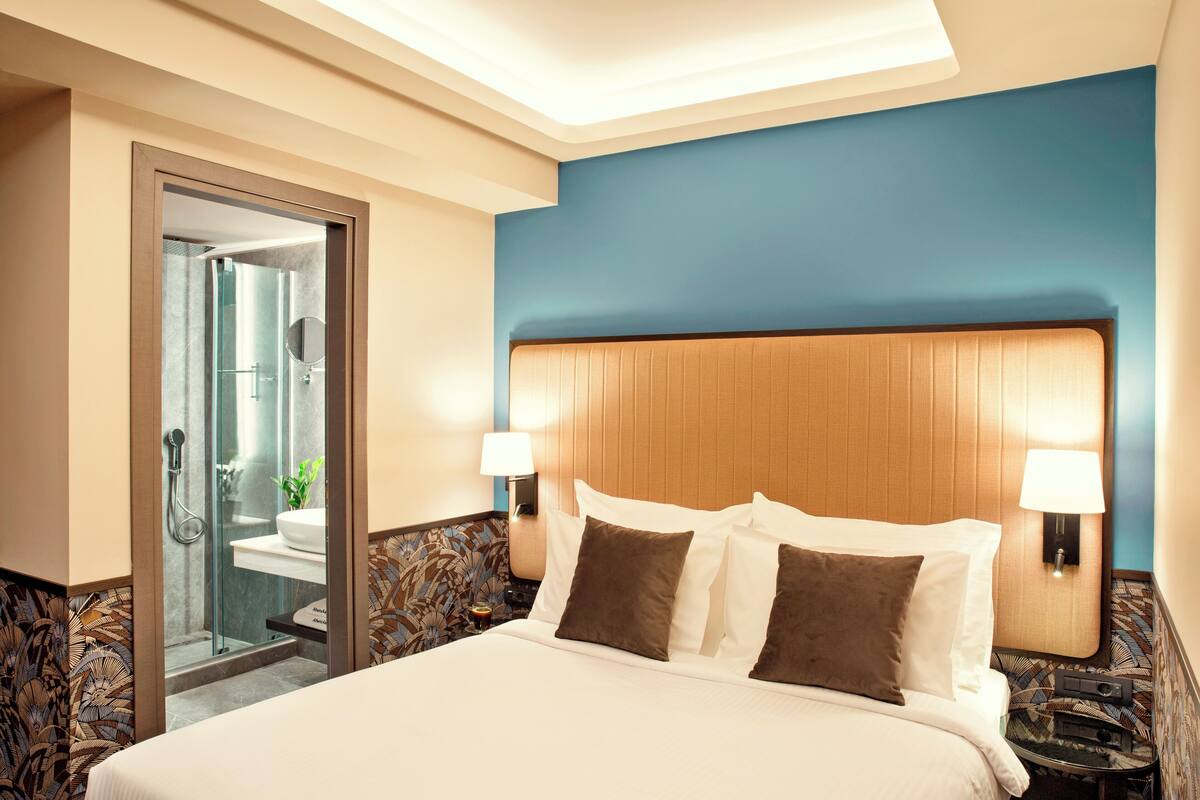 Ein Hotelzimmer mit einem großen Bett, zwei Nachttischlampen, einem hölzernen Kopfteil und einer Duschkabine aus Glas.