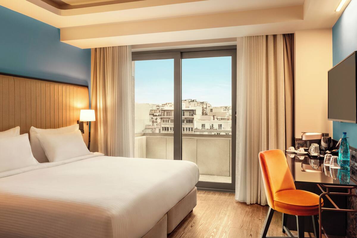Ein Hotelzimmer mit Bett, Schreibtisch mit Stuhl, einem Fenster mit Vorhängen und Blick auf Gebäude.
