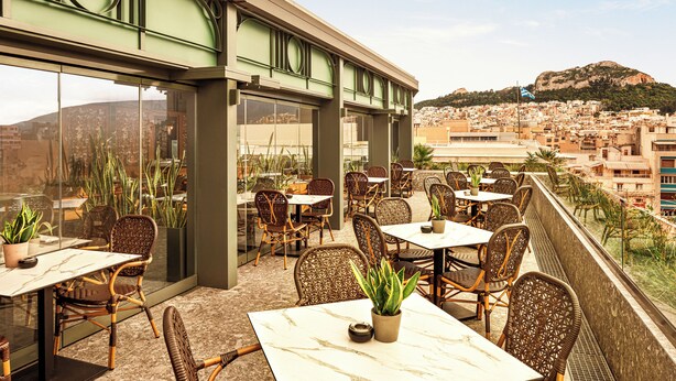 An outdoor dining area with wicker chairs and tables, large windows, and a view of a cityscape and hills. - Zeus Essence Athens Central, Trademark Collection by Wyndham (Athens)