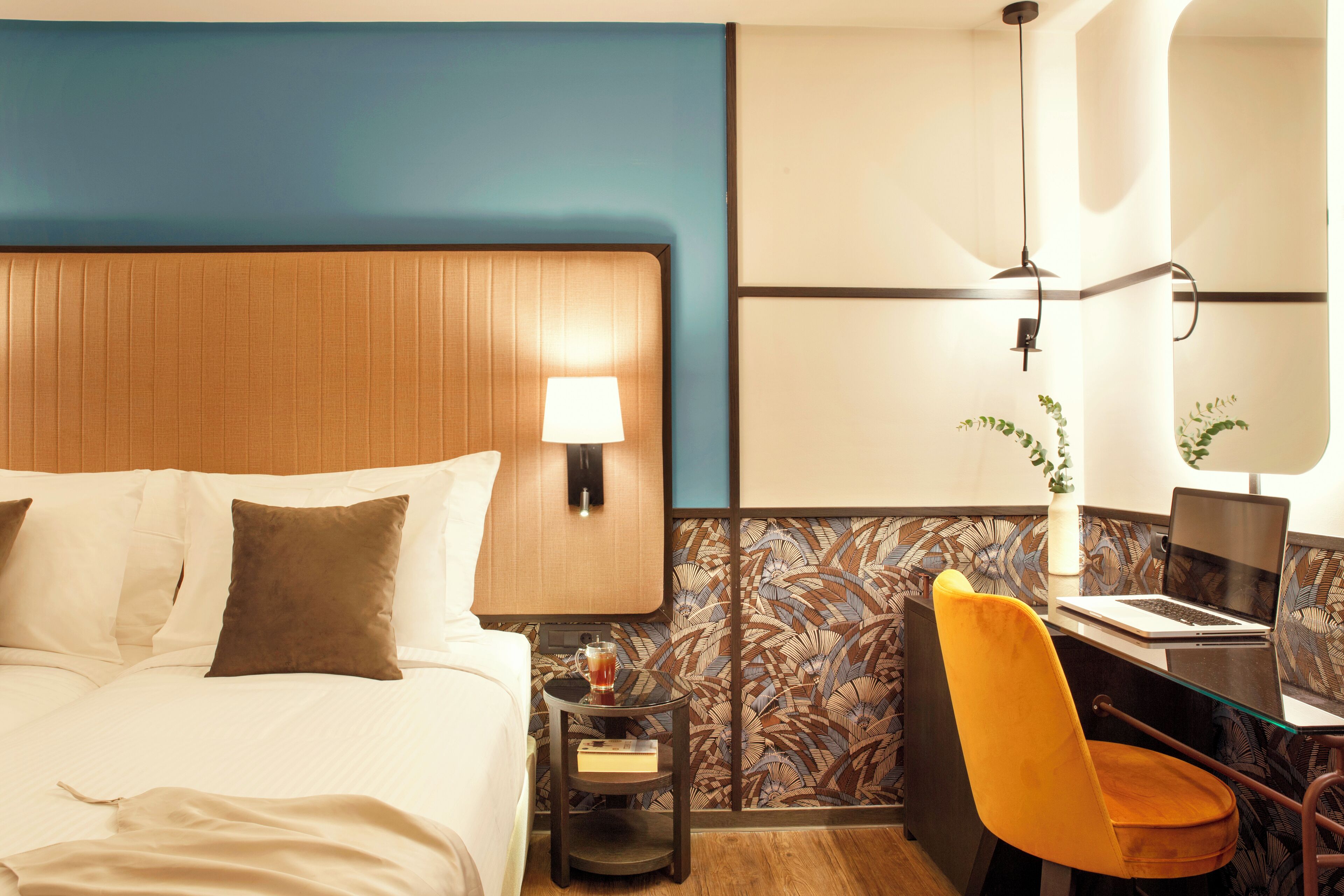 A hotel room with a bed, a desk with a laptop, a chair, a nightstand with a lamp, and a decorative wall panel.