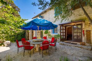 Un bâtiment en pierre à l’aspect rustique, une table ronde entourée de chaises rouges et un parasol bleu offrant de l’ombre.