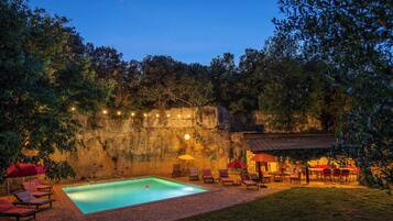 A swimming pool with lounge chairs and umbrellas, surrounded by trees and a stone wall.