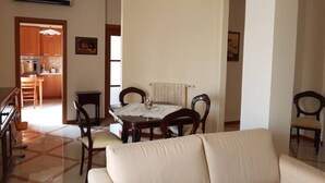 A living room with a beige sofa, a dining table with chairs, and a kitchen area visible through an open doorway.