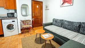 A compact living space with a sofa, a small table, a washing machine, a microwave, and a door with a sign.