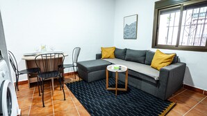 A compact living space with a grey sofa, two yellow cushions, a small round table, a washing machine, and a dining area with chairs and a table. - Marbella Village  | Budget Ground Floor Unit (Marbella)