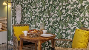 Een eethoek met een tafel gedekt voor twee personen, met rieten stoelen met gele kussens, behang met een patroon en een hangende ijzeren rek.