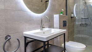 A modern bathroom with a white sink, black metal shelves, a round mirror, and a glass-enclosed shower. - Casa Ancha 10 | Boho Suite (Marbella)