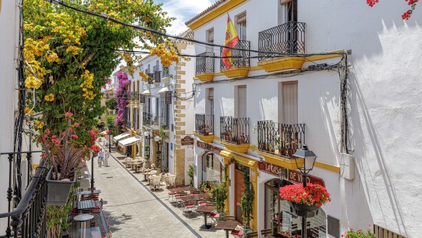A narrow street with outdoor seating, white buildings, balconies, and a flag. - Casa Ancha 10 | Boho Suite (Marbella)