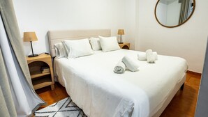 Ein ordentlich bezogenes Bett mit weißen Leinen und Handtüchern, ein runder Spiegel an der Wand und ein Nachttisch aus Holz mit einer Lampe.