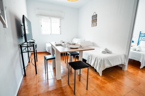 Una habitación limpia y bien iluminada con una mesa de comedor para cuatro personas, un sofá, una cama y un televisor montado en la pared.