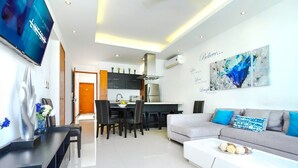 Una sala moderna con sofá, mesa de centro y televisor montado en la pared. Hay un comedor con sillas y una mesa, y una cocina con armarios y estufa.