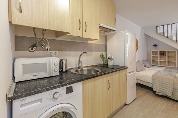 Cocina compacta con lavadora, microondas, lavamanos y una cama en la habitación contigua.