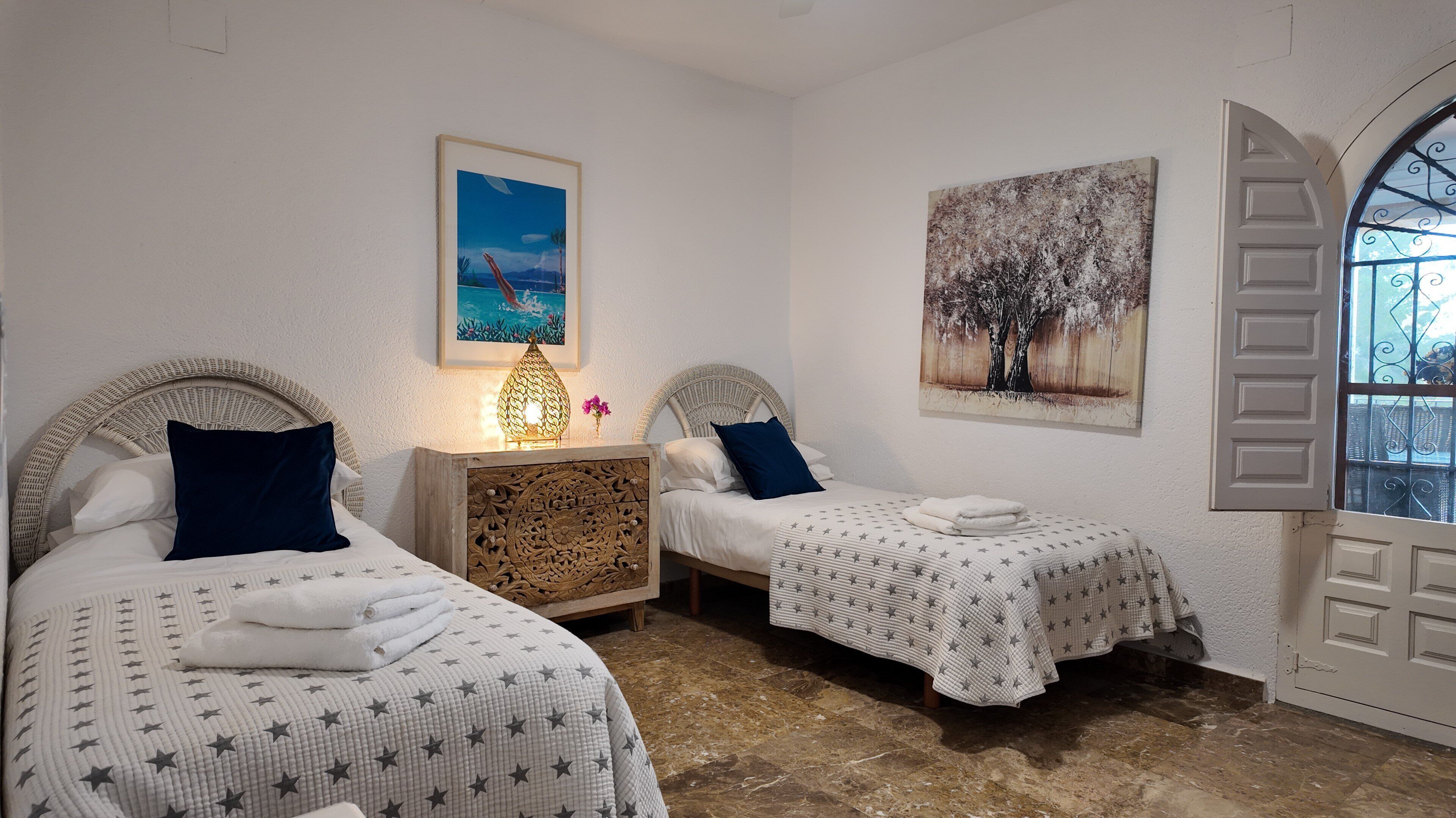 A bedroom with two single beds, a nightstand with a lamp, a painting on the wall, and a window with a decorative door.