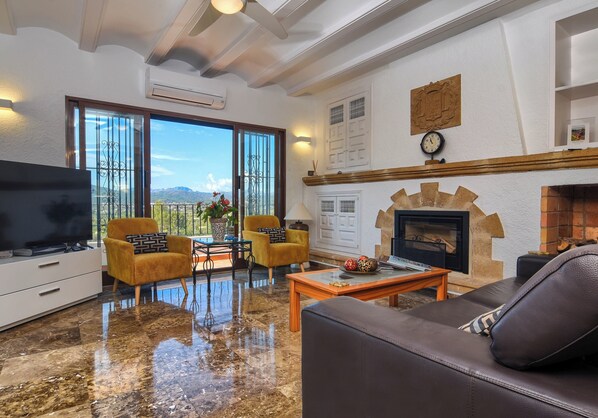 A living room with a fireplace, a sofa, a TV, and a balcony with a view of the mountains. - Sunset Villa Javea with private pool, tennis and padel court (Jávea)