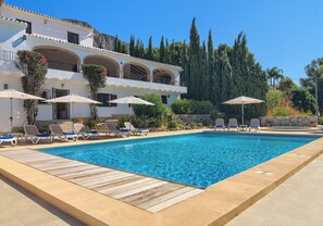 A swimming pool with lounge chairs and umbrellas, surrounded by a building with balconies and cypress trees. - Sunset Villa Javea with private pool, tennis and padel court (Jávea)