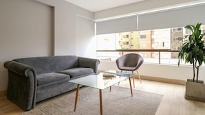 Ett modernt vardagsrum med en grå soffa, ett soffbord i glas och en grå fåtölj. Ett stort fönster ger utsikt över byggnaderna utanför.