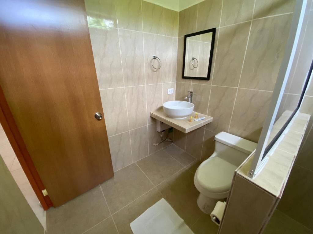 Un baño con puerta de madera, paredes de azulejos color beige, un lavamanos blanco, un inodoro y un espejo.