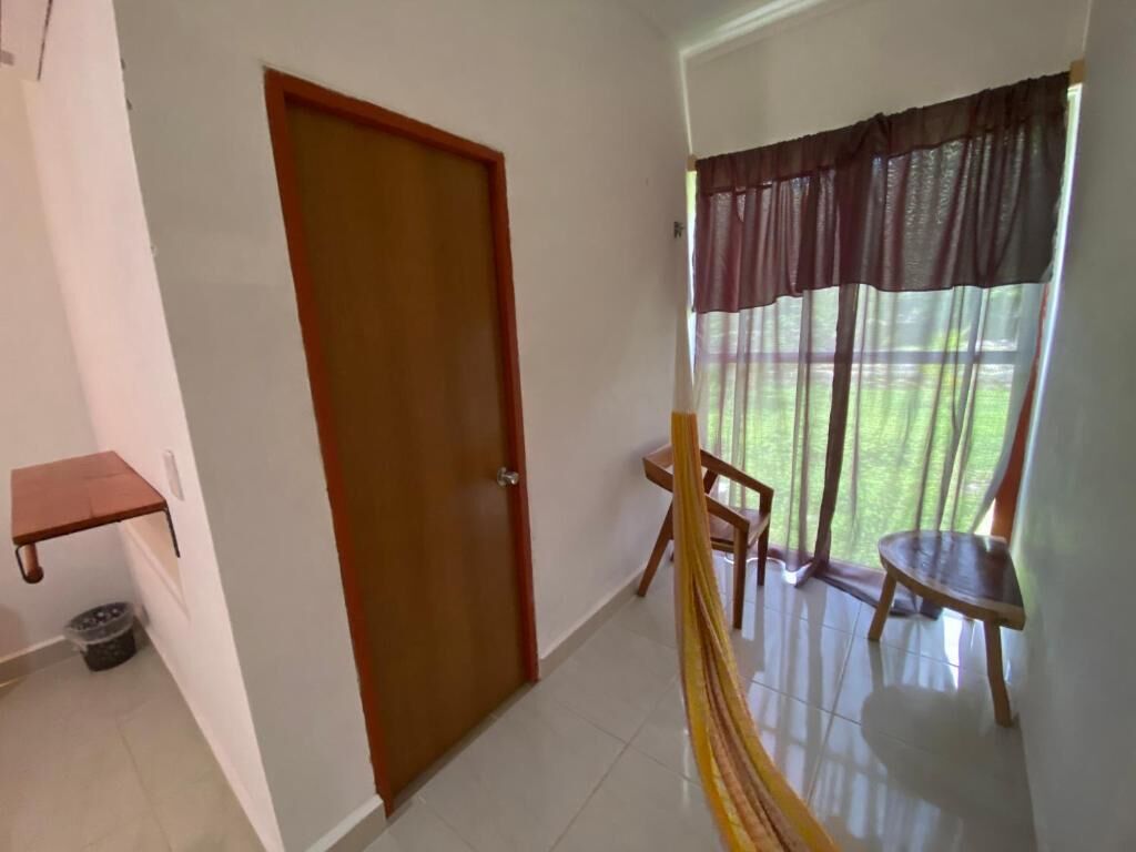 A hallway with a wooden door, a small shelf, a chair, and a view of a grassy area through a window with a curtain.