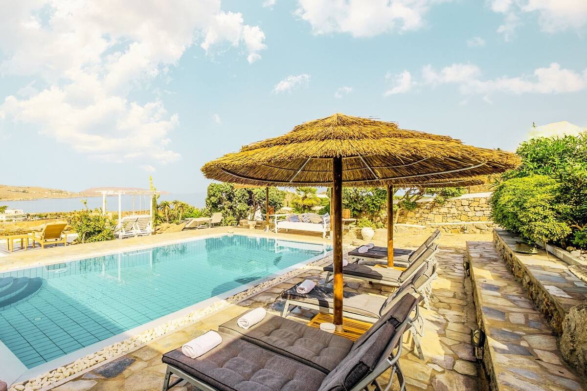 Zona de piscina con sillas de descanso y una sombrilla de paja, rodeada de muros de piedra y vegetación.