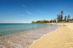 A beach with clear water, a boat on the sand, and buildings in the background.