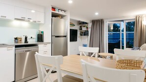 A modern kitchen with white cabinets, a stainless steel refrigerator, and a dishwasher. There is a dining area with a wooden table and white chairs. - Moffat Beach Coastal Contemporary Unit B (Moffat Beach)
