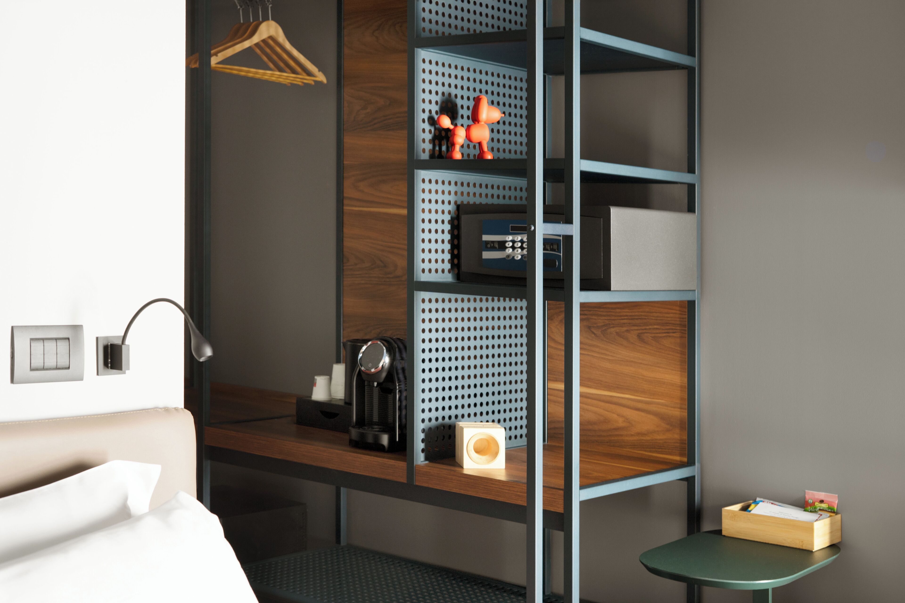 Camera d'albergo con un letto, una scrivania con una macchina per il caffè, uno scaffale con oggetti decorativi e un piccolo tavolino con una scatola.