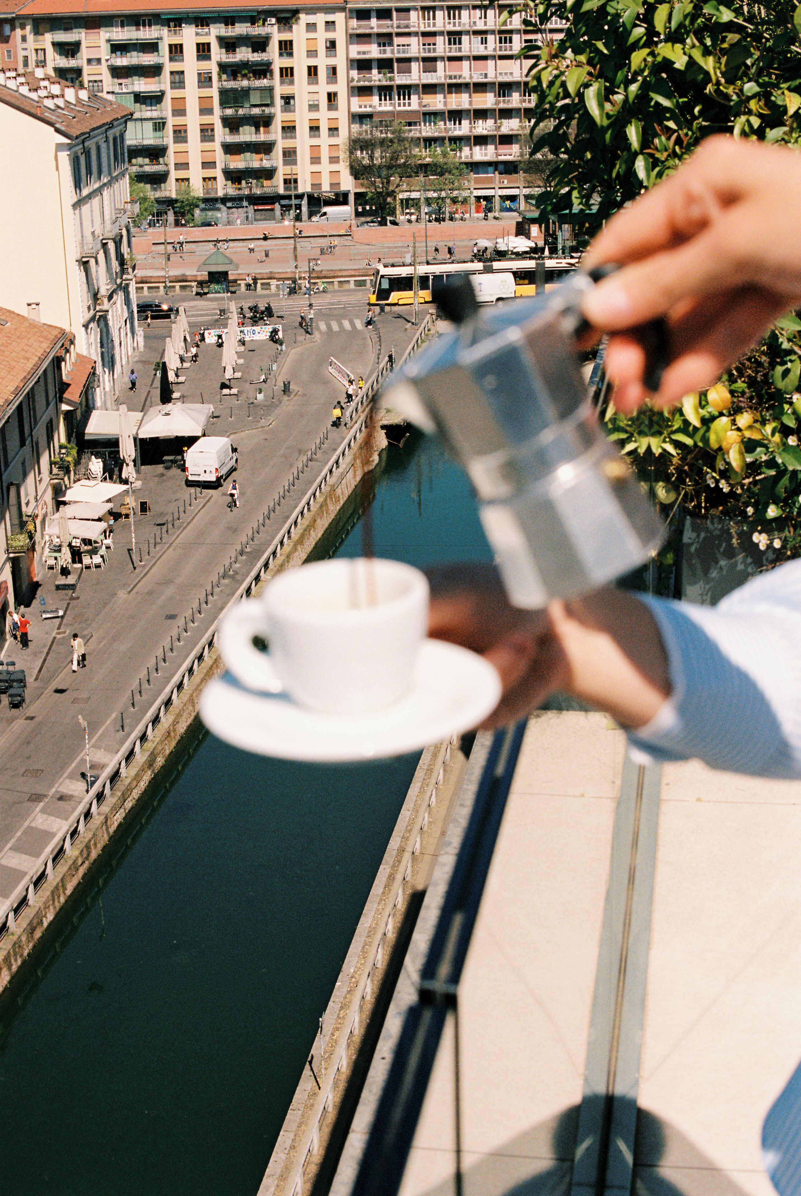 A person pouring coffee into a cup on a balcony overlooking a canal with buildings and a street.