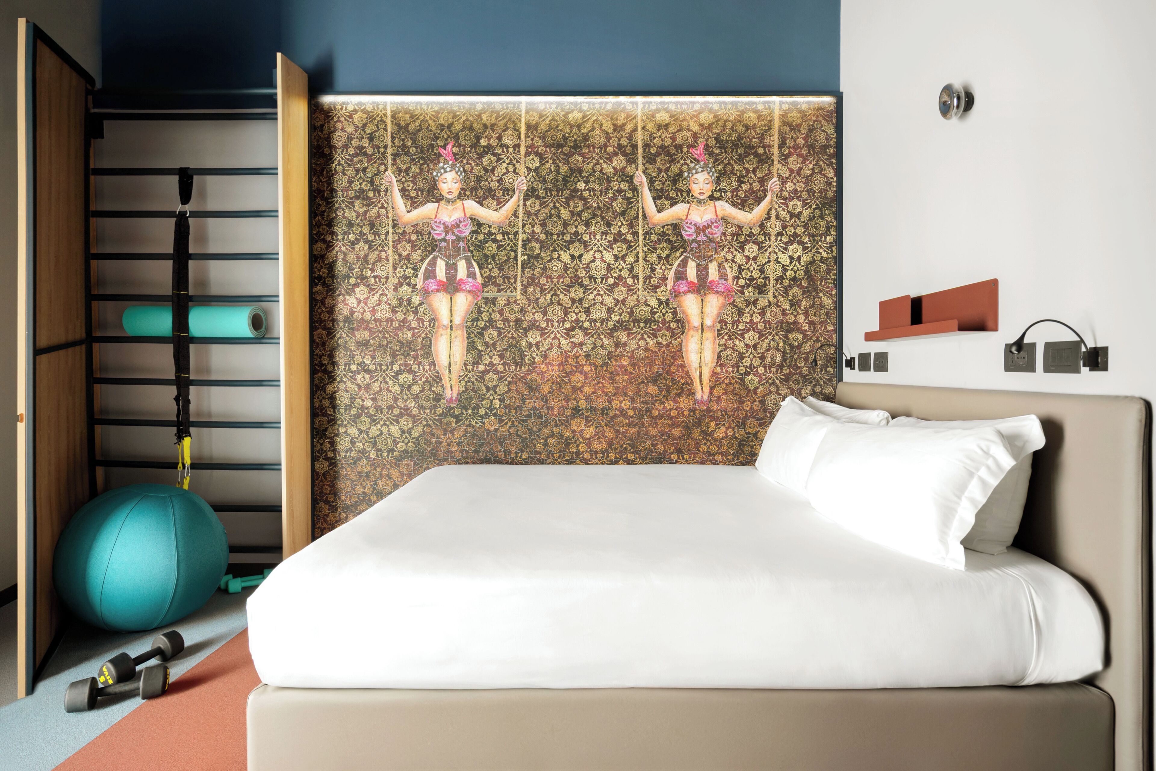 Ein Hotelzimmer mit Bett, Gymnastikball, Kurzhanteln, einer Wandablage und einem gemusterten Kopfteil.